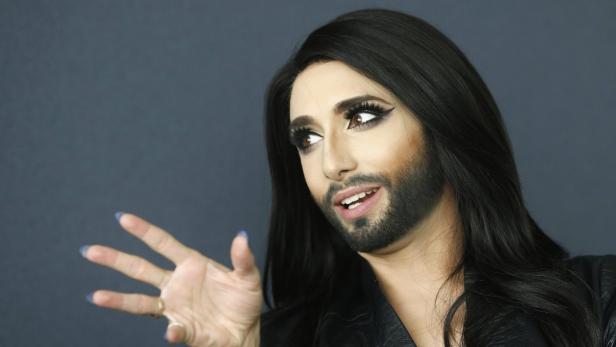 Conchita Wurst mit dunklem Haar und Bart gestikuliert vor einem grauen Hintergrund.