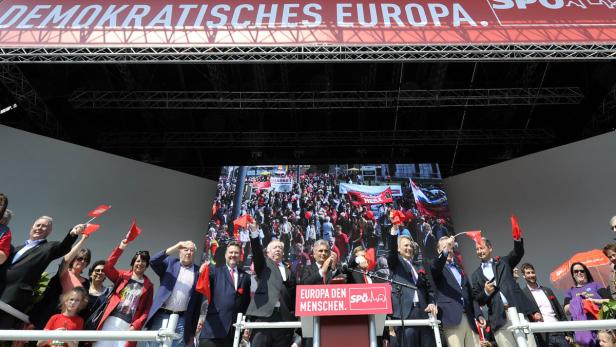 Eine Gruppe von Politikern der SPÖ winkt auf einer Bühne mit roten Fahnen.