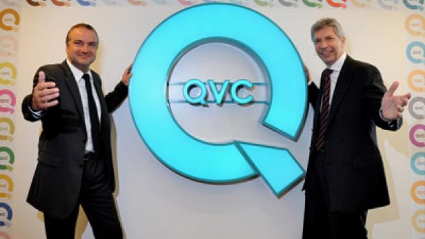 Zwei Männer in Anzügen posieren neben einem großen, leuchtenden QVC-Logo.