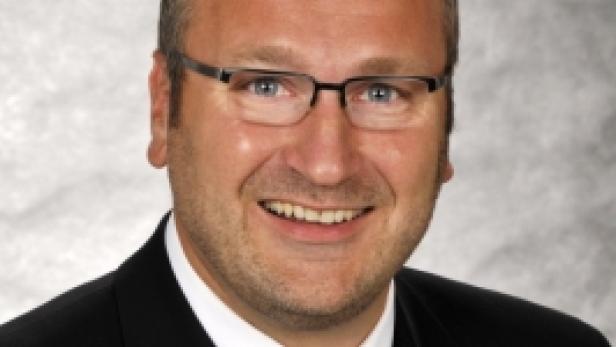 Ein lächelnder Mann mit Brille und Anzug vor einem grauen Hintergrund.