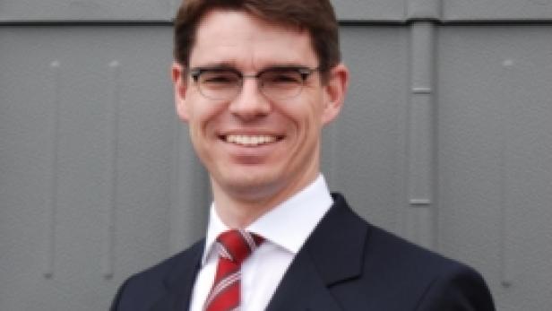 Ein lächelnder Mann mit Brille, Anzug und Krawatte vor einer grauen Wand.