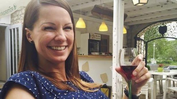 Eine Frau lächelt und hält ein Glas Rotwein in einem Außenbereich.