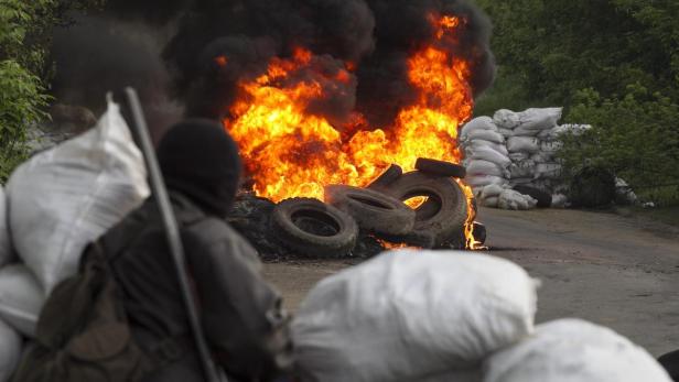 Ein vermummter Mann steht vor einem Reifenstapel, der in Flammen steht, und Sandsäcken.