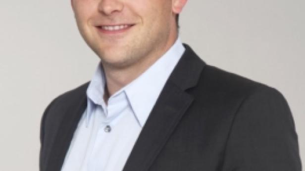 Ein lächelnder Mann in einem dunklen Anzug und hellblauem Hemd vor einem grauen Hintergrund.