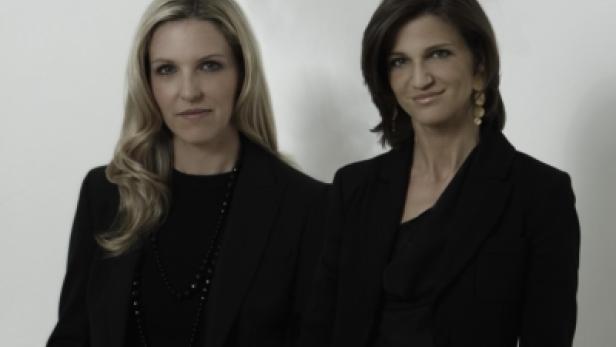 Zwei Frauen in schwarzen Blazern stehen nebeneinander vor einem hellen Hintergrund.