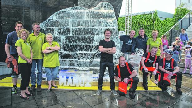 Eine Gruppe von Menschen posiert vor einer Eisstatue mit der Aufschrift „MAMUZ“.
