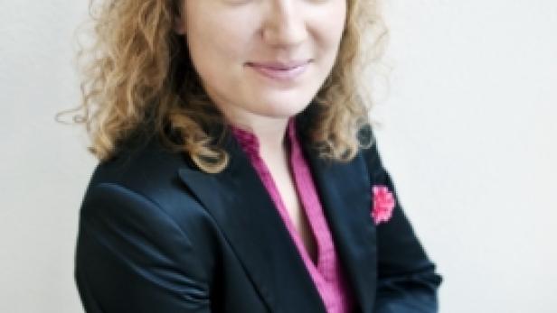 Eine Frau mit lockigem Haar trägt einen Blazer und einen auffälligen Ring.