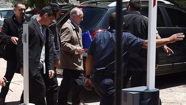 Ein älterer Mann mit brauner Jacke und blauem Ordner wird von Sicherheitsleuten begleitet.