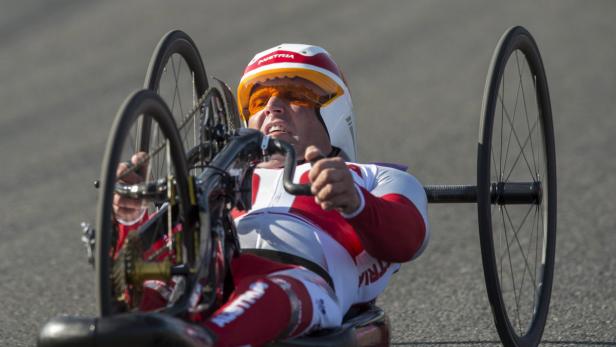 Ein Handbiker in rot-weißer Sportkleidung und Helm fährt auf einer asphaltierten Straße.