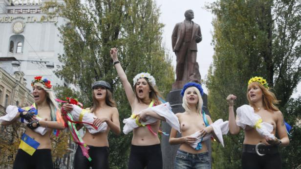 Fünf barbusige Frauen mit Blumenkränzen protestieren vor einem Lenin-Denkmal.