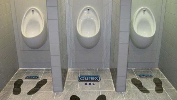 In einer Herrentoilette weisen Fußabdrücke auf dem Boden zu Urinalen mit Durex-Werbung.