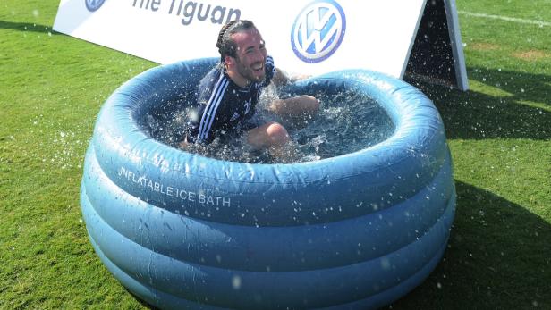 Ein Mann sitzt lachend in einem aufblasbaren Eisbad auf einer Wiese.