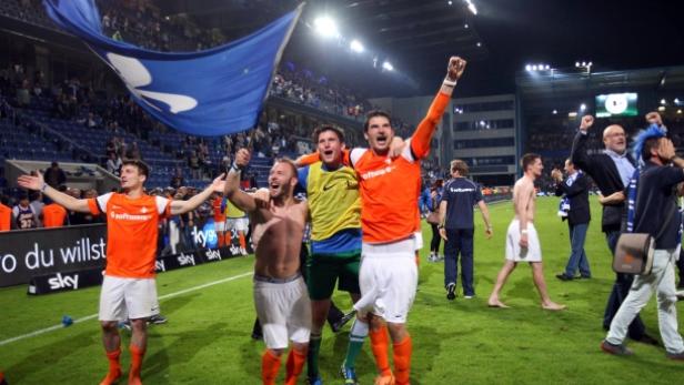 Mehrere Fußballspieler feiern auf dem Rasen mit erhobenen Armen und einem blauen Banner.