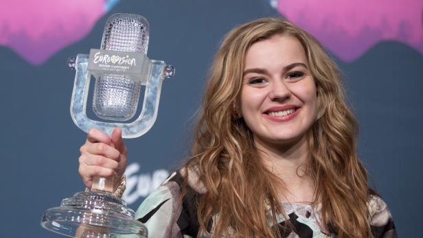 Eine junge Frau hält eine Eurovision-Trophäe in die Höhe und lächelt.