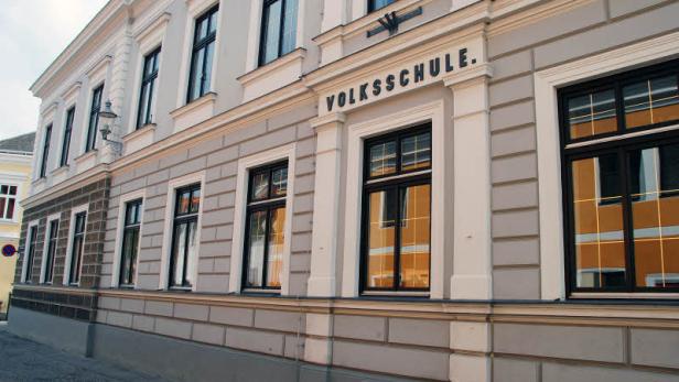 Die Fassade einer hellgrauen Volksschule mit vielen Fenstern.