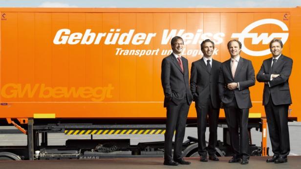 Vier Männer in Anzügen posieren vor einem orangefarbenen LKW von Gebrüder Weiss.