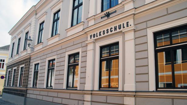 Die Fassade einer Volksschule mit großen Fenstern und dem Schriftzug „Volksschule“.