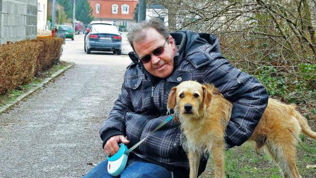 Ein Mann mit Sonnenbrille sitzt mit seinem hellbraunen Hund auf einer Bank am Straßenrand.