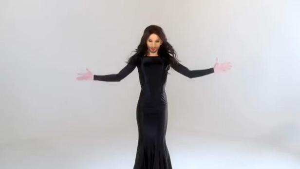 Eine Frau mit Bart und Make-up trägt ein langes, schwarzes Kleid und streckt die Arme aus.