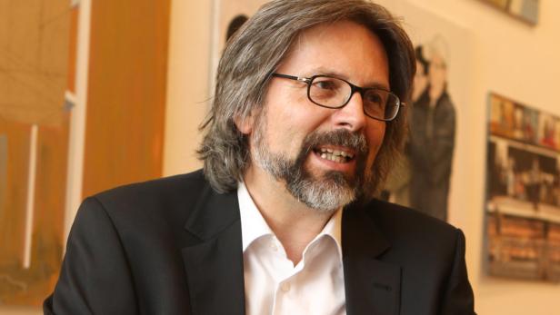 Ein Mann mit Brille, Bart und dunklem Anzug lächelt.