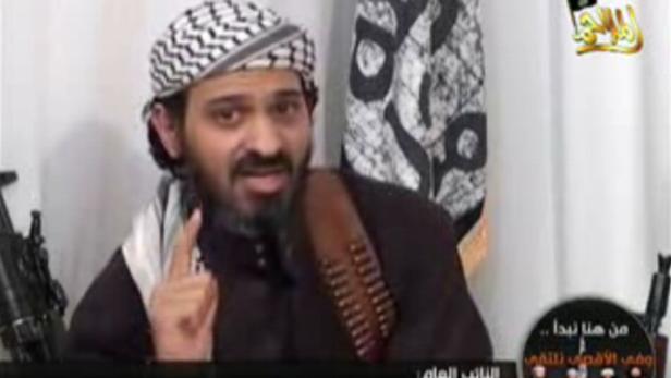 Ein Mann mit Turban und Munitionsgürtel gestikuliert vor einer Flagge.