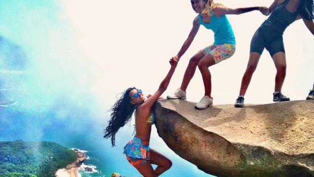 Eine Frau wird von zwei Freundinnen auf einem Felsen über einer Küstenlandschaft gehalten.