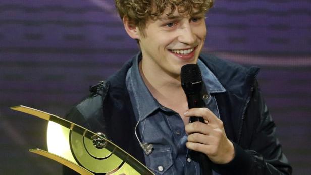 Ein lächelnder junger Mann hält eine goldene Auszeichnung und ein Mikrofon.