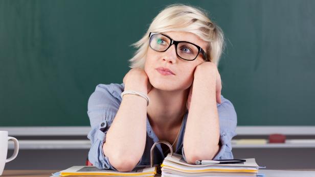 Eine Lehrerin sitzt nachdenklich vor einer Tafel und einem Stapel Papier.
