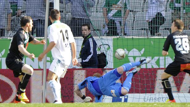 Ein Fußballtorwart hechtet nach dem Ball, während zwei Spieler zusehen.
