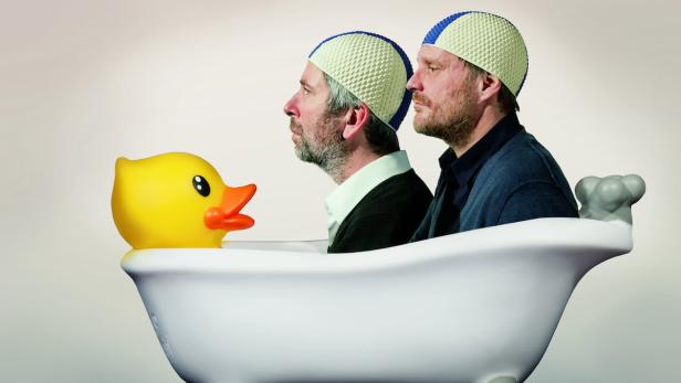 Zwei Männer mit Badekappen sitzen mit einer gelben Gummiente in einer Badewanne.