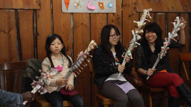 Drei Frauen sitzen auf Stühlen und halten Papierstäbe in einem Raum mit Holzwänden.