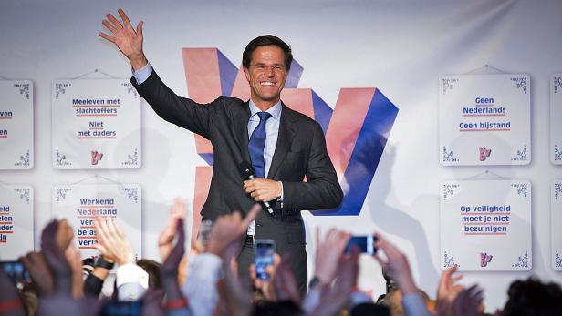 Mark Rutte winkt bei einer Wahlkampfveranstaltung vor einer Wand mit VVD-Slogans.