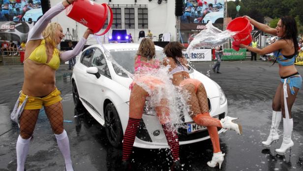 Mehrere Frauen in knapper Bekleidung waschen ein weißes Auto mit Wasser aus Eimern.
