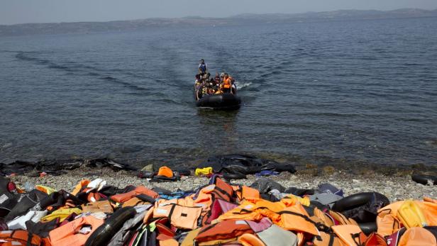 Ein Schlauchboot mit Menschen nähert sich einem Strand, an dem viele Schwimmwesten liegen.