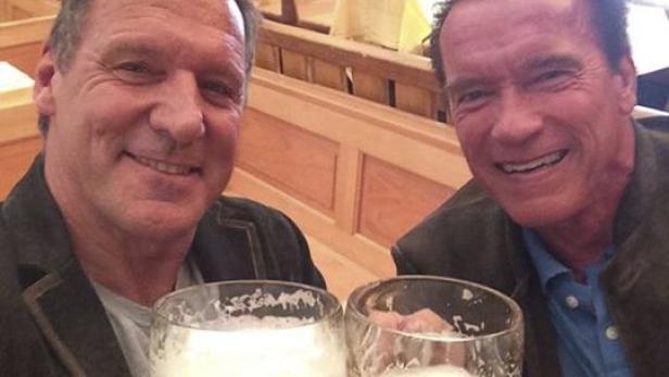 Arnold Schwarzenegger und ein Mann stoßen mit Bierkrügen an.