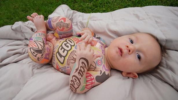 Ein Baby liegt auf einer Decke, dessen Körper mit Logos von Coca-Cola, Lego und anderen Marken bedeckt ist.