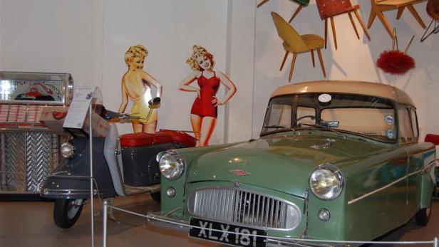 Ein grünes Auto und ein grauer Motorroller stehen vor zwei Aufstellern von Frauen in Badeanzügen.