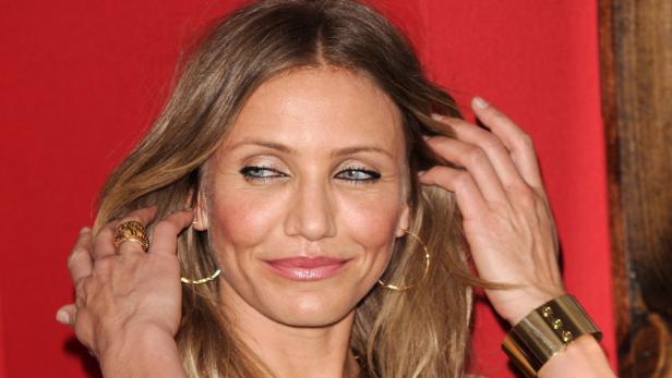 Cameron Diaz posiert mit goldenem Armreif und Ohrringen vor einem roten Hintergrund.