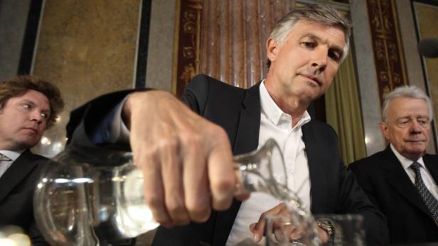 Ein Mann schenkt Wasser aus einer Karaffe in ein Glas ein, während zwei weitere Männer zusehen.