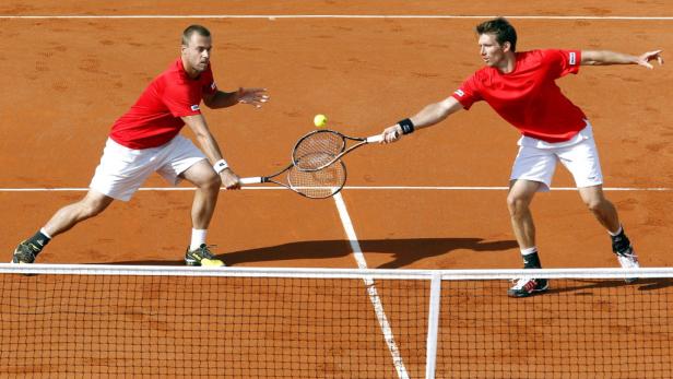 Zwei Tennisspieler in roten Trikots spielen ein Doppel auf einem Sandplatz.