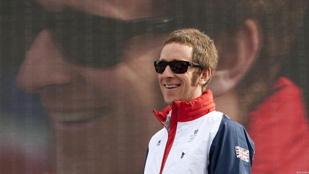 Bradley Wiggins mit Sonnenbrille und britischer Olympia-Jacke vor einer großen Leinwand.