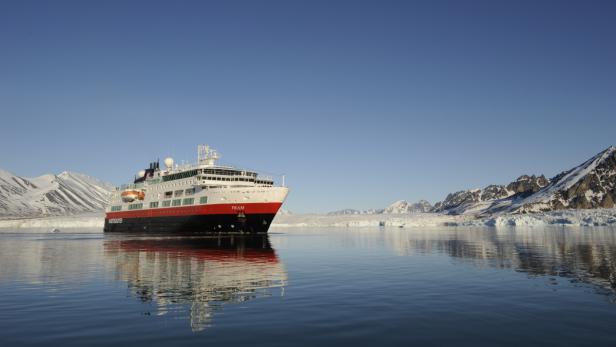 Das Schiff „Fram“ fährt durch ruhiges Wasser vor einer schneebedeckten Landschaft.