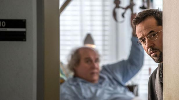 Ein Mann mit Brille späht um eine Ecke, während im Hintergrund eine Person im Krankenbett liegt.