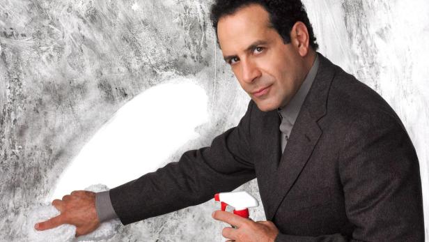 Adrian Monk putzt mit einem Lappen und einer Sprühflasche eine Wand.