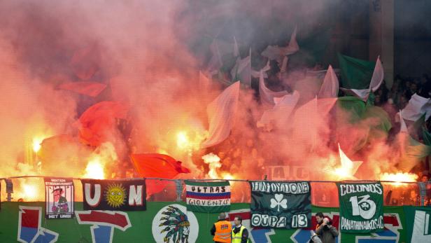 Fußballfans zünden Bengalos und schwenken Fahnen hinter einem Zaun.
