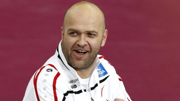 Ein Mann mit Glatze lächelt in einer weißen Jacke vor einem roten Hintergrund.