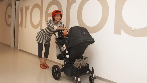 Eine Frau mit rotem Hut und Kinderwagen steht vor einer Wand mit der Aufschrift „Herren Garderobe“.