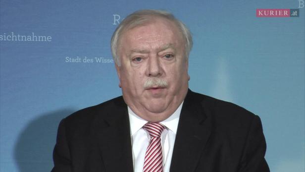 Ein Mann mit Schnurrbart und Anzug vor einem blauen Hintergrund.