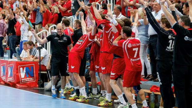 Ein Handballtrainer und seine Mannschaft jubeln am Spielfeldrand.