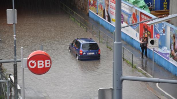 Ein blaues Auto fährt durch eine überflutete Unterführung; ein ÖBB-Schild ist im Vordergrund zu sehen.